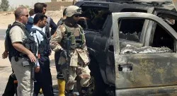 A private security contractor (L) and soldiers look at a destroyed vehicle after an attack near the Iraqi city of Najaf May 18, 2006. Witnesses said a roadside bomb exploded near a convoy of vehicles commonly used by private security firms, killing one Iraqi policeman. REUTERS/Ali Abu Shish - RTR1DHVM